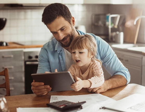 Niña pequeña utilizando una tablet sentada encima de su padre en la cocina de su casa